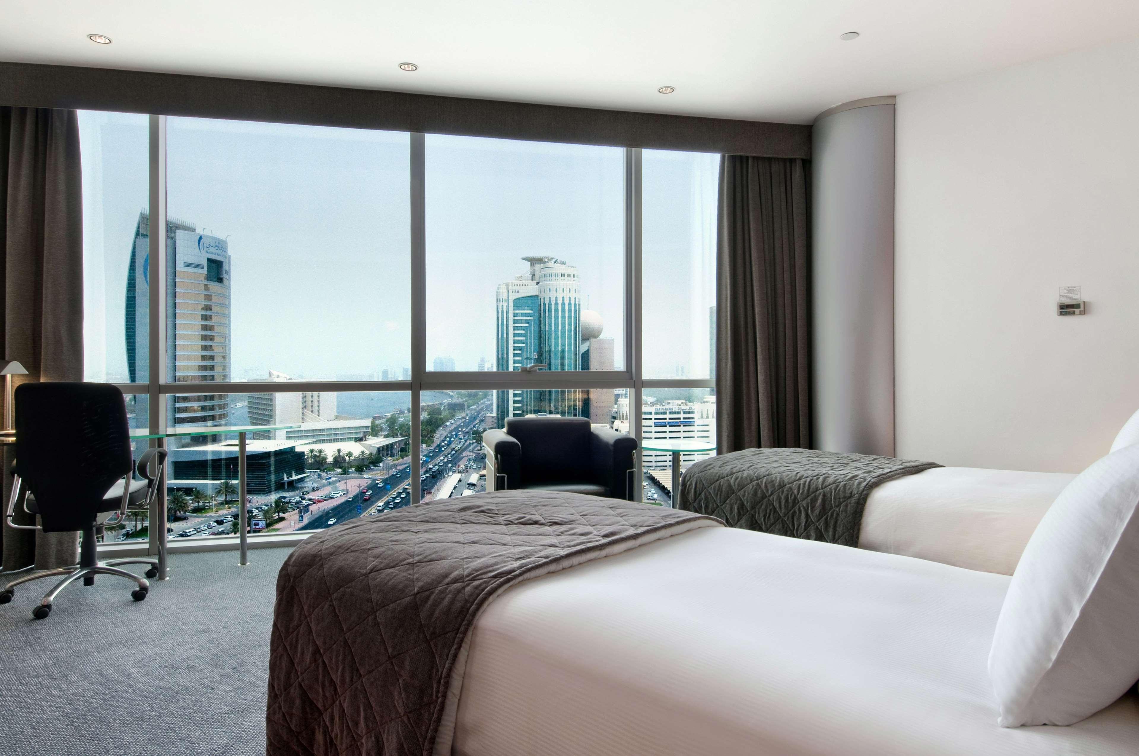 Golden Sands Boutique Hotel-Dubai Creek Стая снимка The photo shows a modern hotel room with two beds. The beds are neatly made with white linens and dark gray throws. There is a large window that offers a view of a cityscape, featuring tall buildings and skyscrapers. The room has contemporary furnish