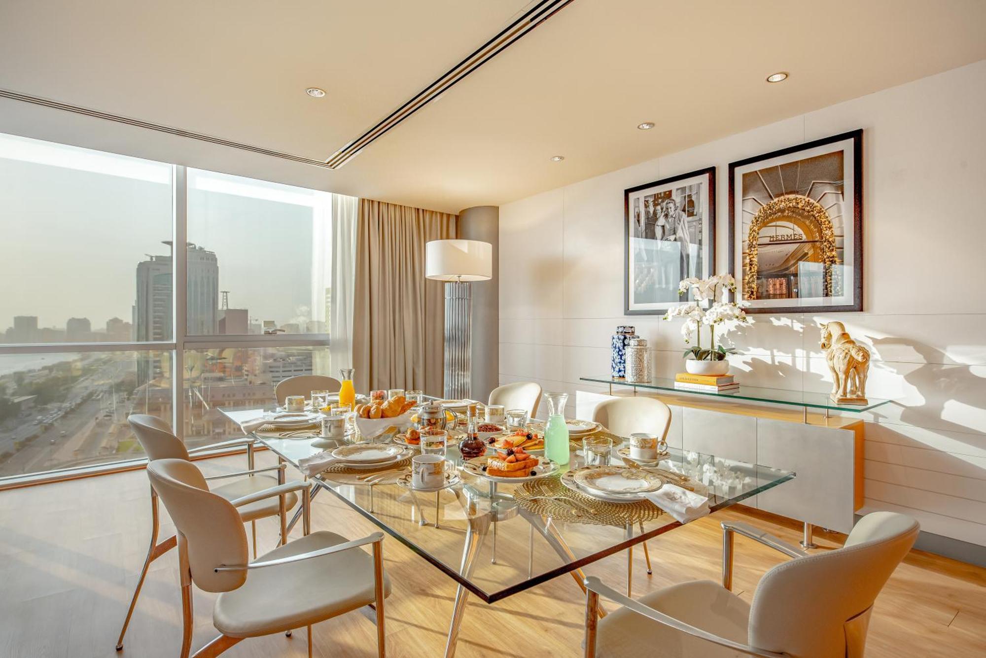 Golden Sands Boutique Hotel-Dubai Creek Стая снимка The photo shows a modern dining room featuring a glass dining table set for a meal. The table is adorned with plates, cutlery, glasses, and a variety of food items, suggesting a breakfast or brunch setting. Surrounding the table are sleek, white chai