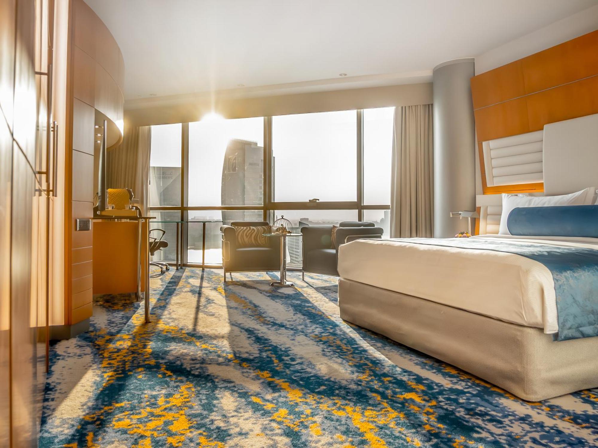 Golden Sands Boutique Hotel-Dubai Creek Стая снимка The photo shows a modern hotel room featuring a large bed with a light beige cover and a blue accent blanket. Large windows allow natural light to stream in, revealing a scenic view outside. The floor is adorned with a colorful area rug in shades of 