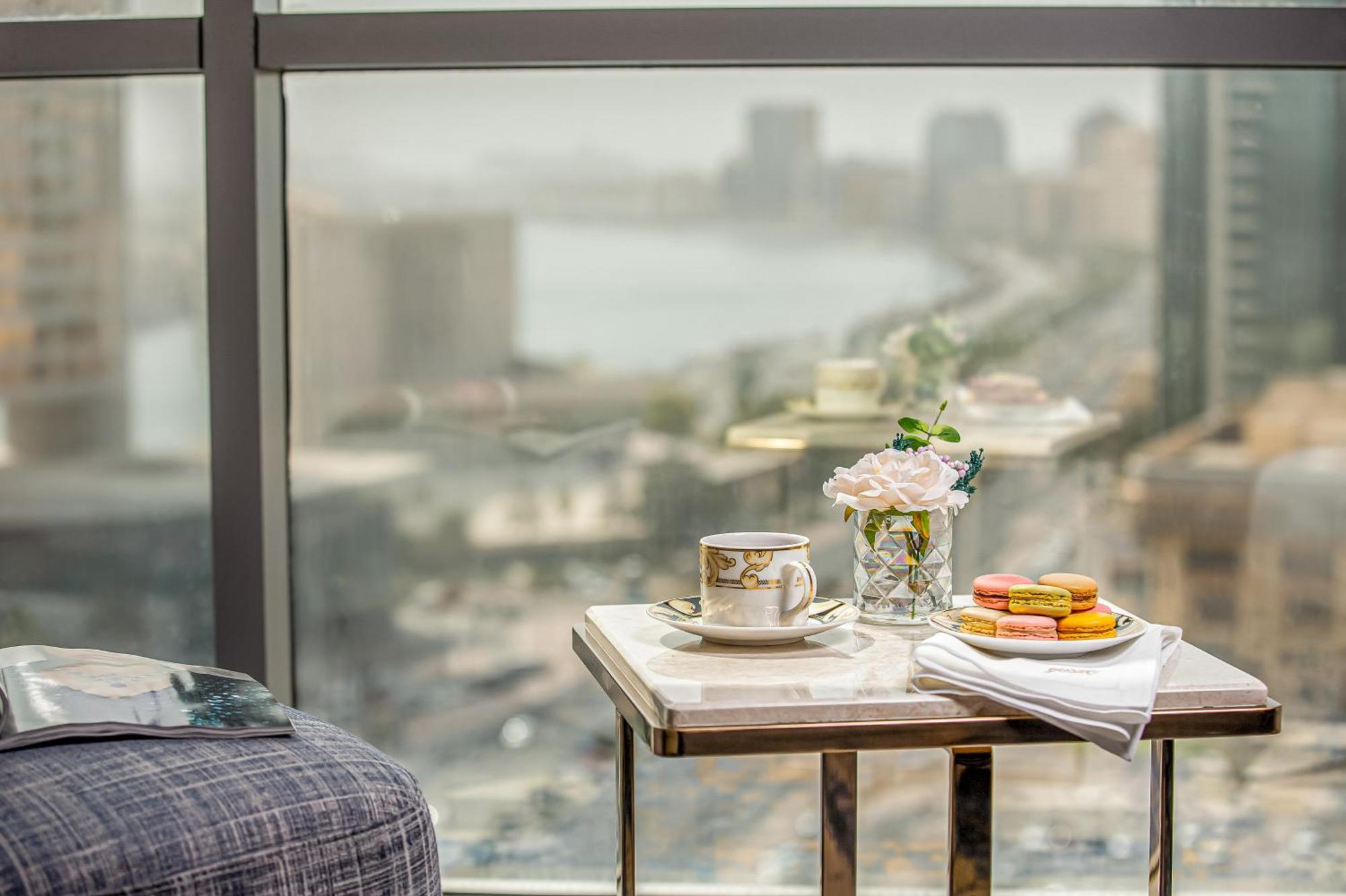 Golden Sands Boutique Hotel-Dubai Creek Екстериор снимка The photo shows a cozy setting with a small table placed next to a large window. On the table, there is a fine china cup with a saucer, a glass vase containing fresh flowers, and a plate of colorful macarons. A neatly folded napkin is also present. T