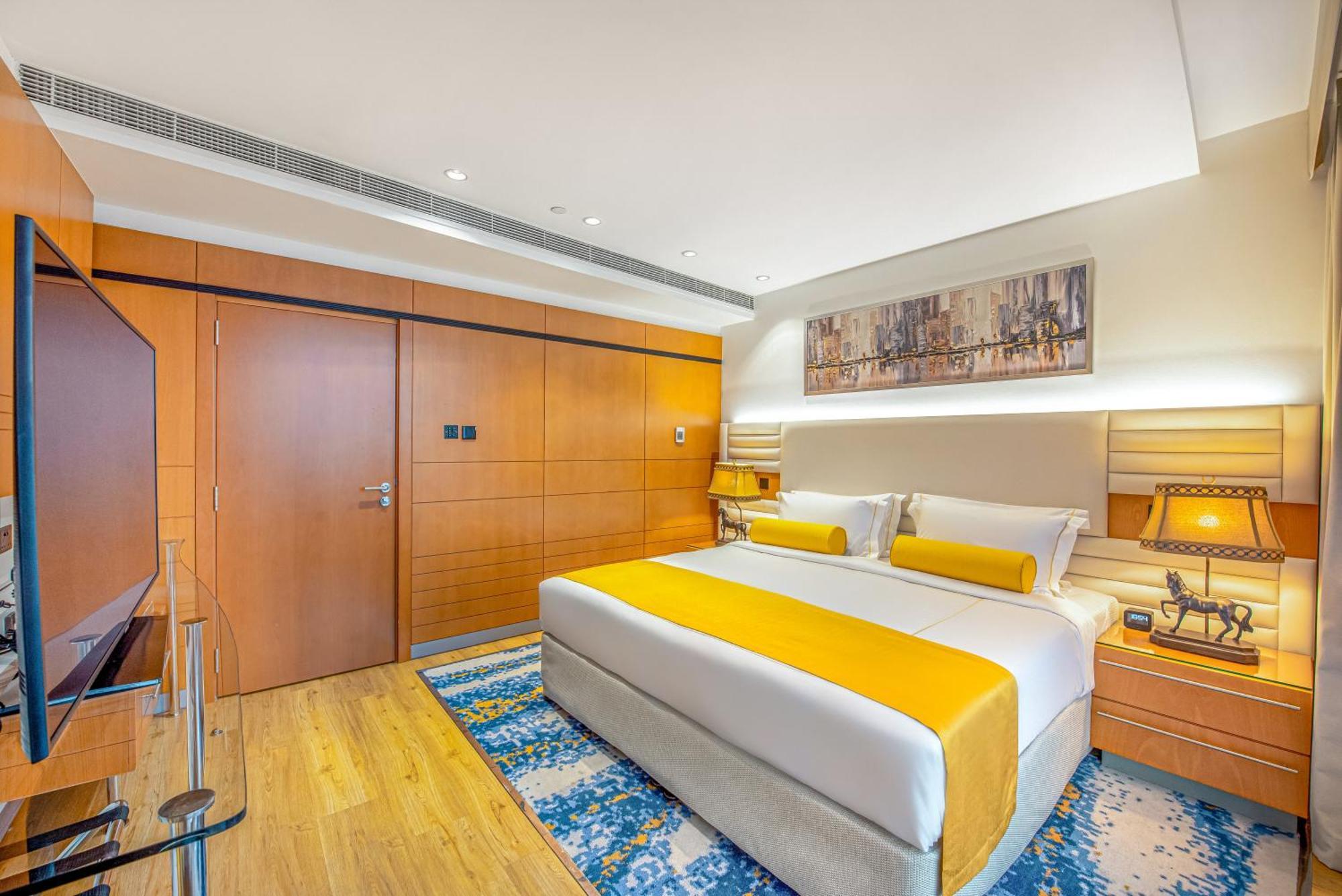 Golden Sands Boutique Hotel-Dubai Creek Стая снимка The photo shows a modern hotel room featuring a king-sized bed with bright yellow accents on the bedding. The walls are a warm wood tone, with a large piece of artwork above the bed. There are nightstands on either side of the bed, each with lamps. A