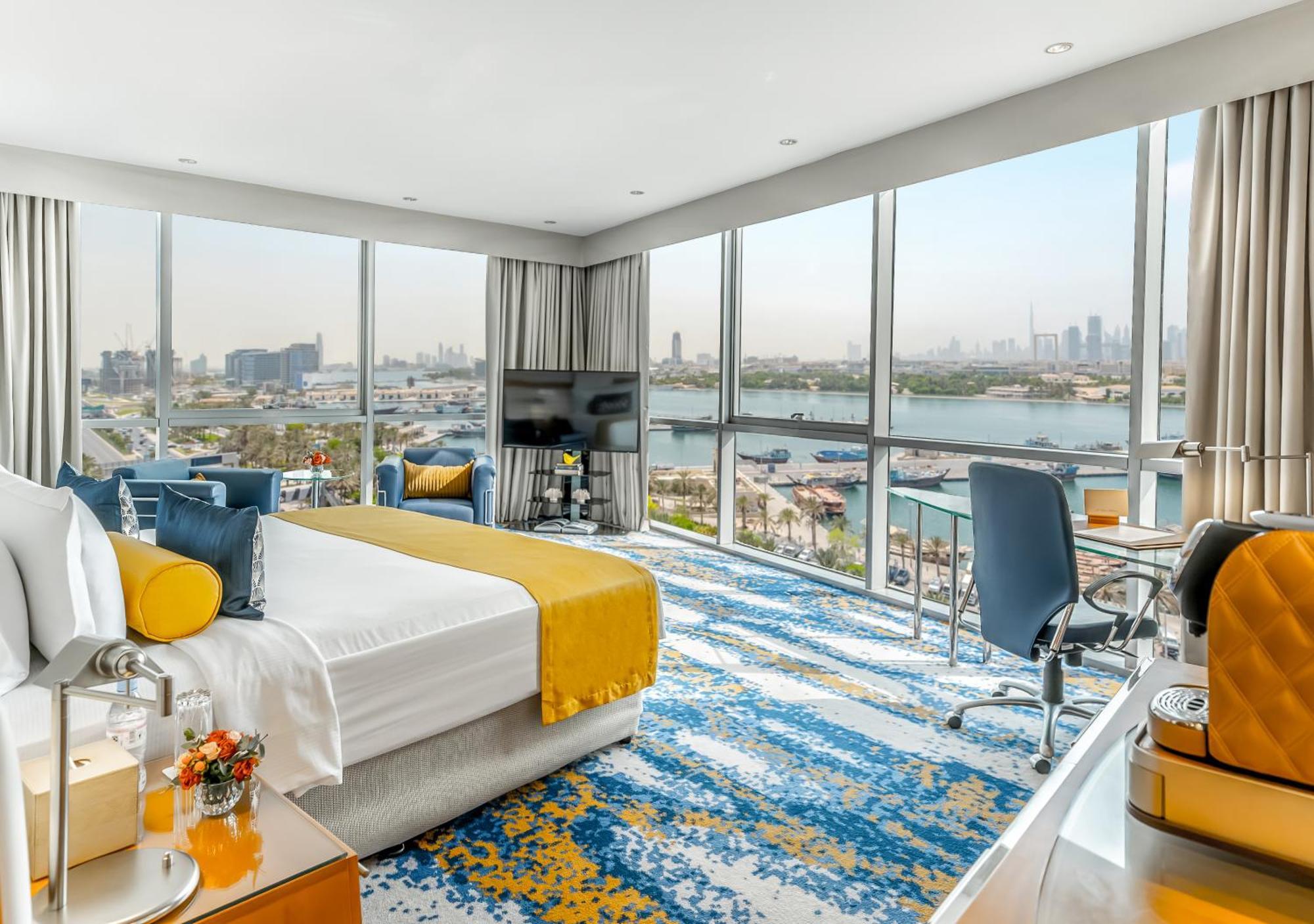 Golden Sands Boutique Hotel-Dubai Creek Екстериор снимка The photo shows a modern hotel room with a spacious layout and large windows that offer a view of the city and water. The room features a comfortable bed with a bright yellow and white duvet, a stylish area rug with a blue and yellow pattern, and con