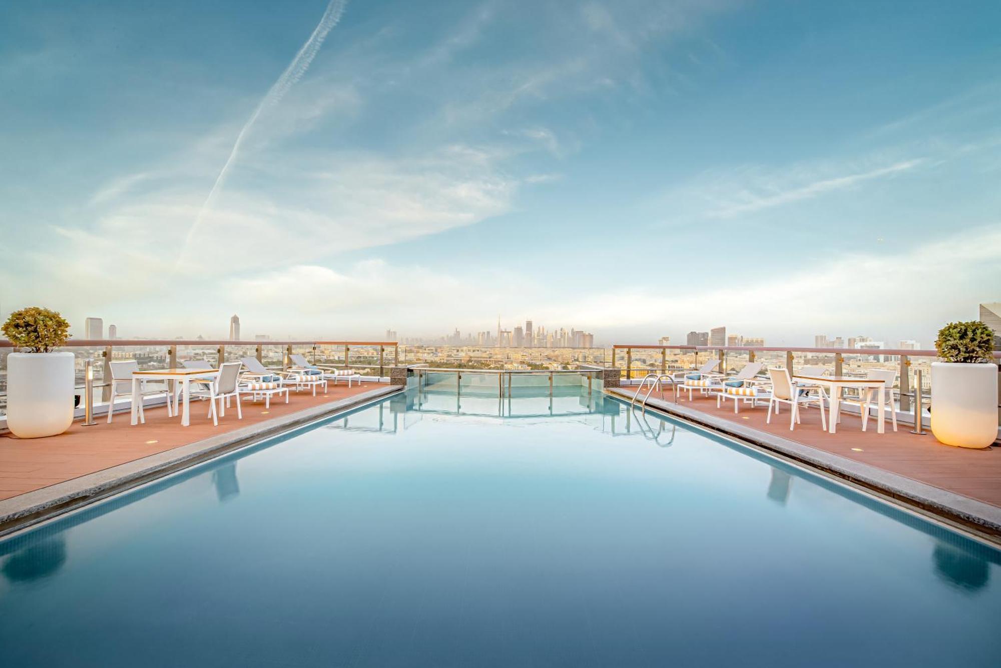 Golden Sands Boutique Hotel-Dubai Creek Екстериор снимка The photo showcases a rooftop swimming pool with a stunning view of a city skyline in the background. The pool is calm and reflects the sky, which is clear with a few clouds. Surrounding the pool are several white lounge chairs arranged for relaxatio