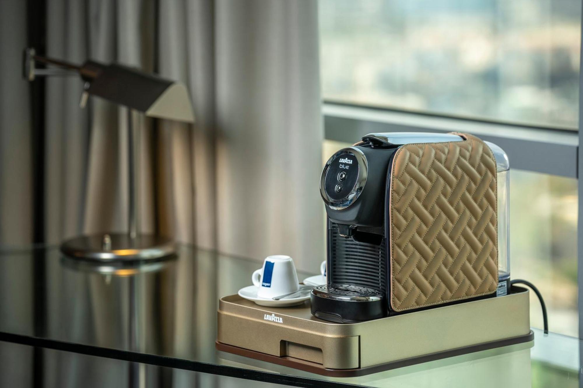 Golden Sands Boutique Hotel-Dubai Creek Стая снимка The photo features a coffee machine placed on a sleek table. The machine has a stylish design with a patterned brown exterior. Next to the machine, there is a small saucer with a blue and white coffee cup. The background includes a large window with 