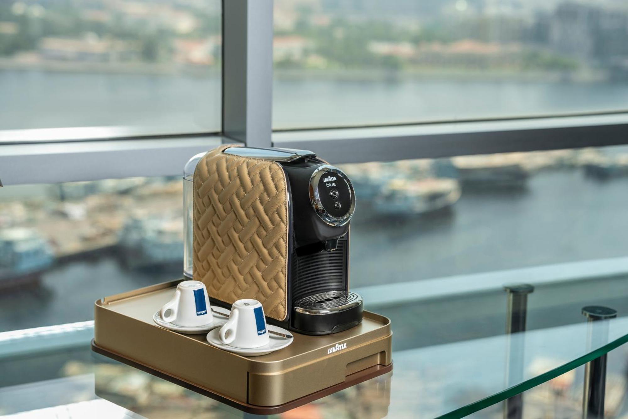 Golden Sands Boutique Hotel-Dubai Creek Екстериор снимка The image shows a coffee machine placed on a glass table with a scenic view of water in the background. The coffee machine has a sleek design, featuring a beige and black color scheme. There are two coffee cups positioned next to the machine, along w